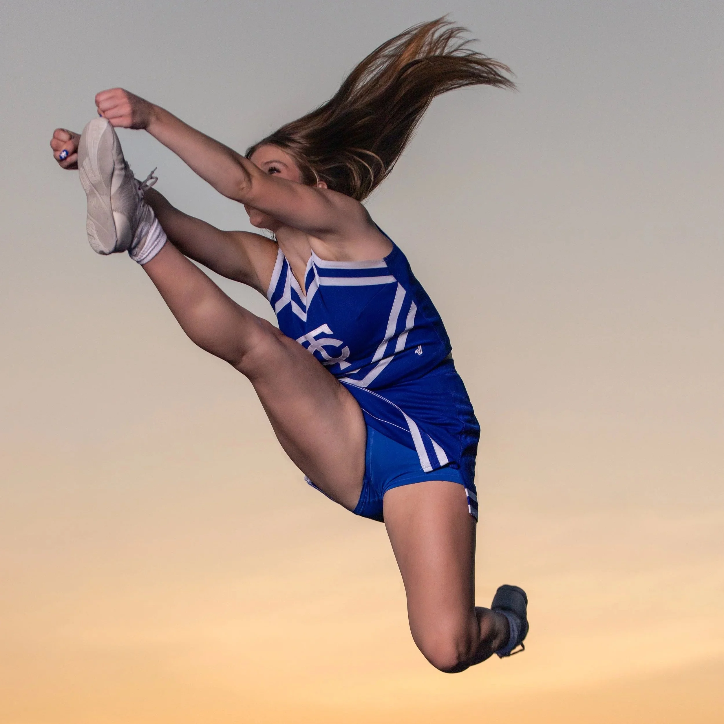 A cheerleader in a blue uniform mid-jump with her legs extended in a toe touch against a warm sky.