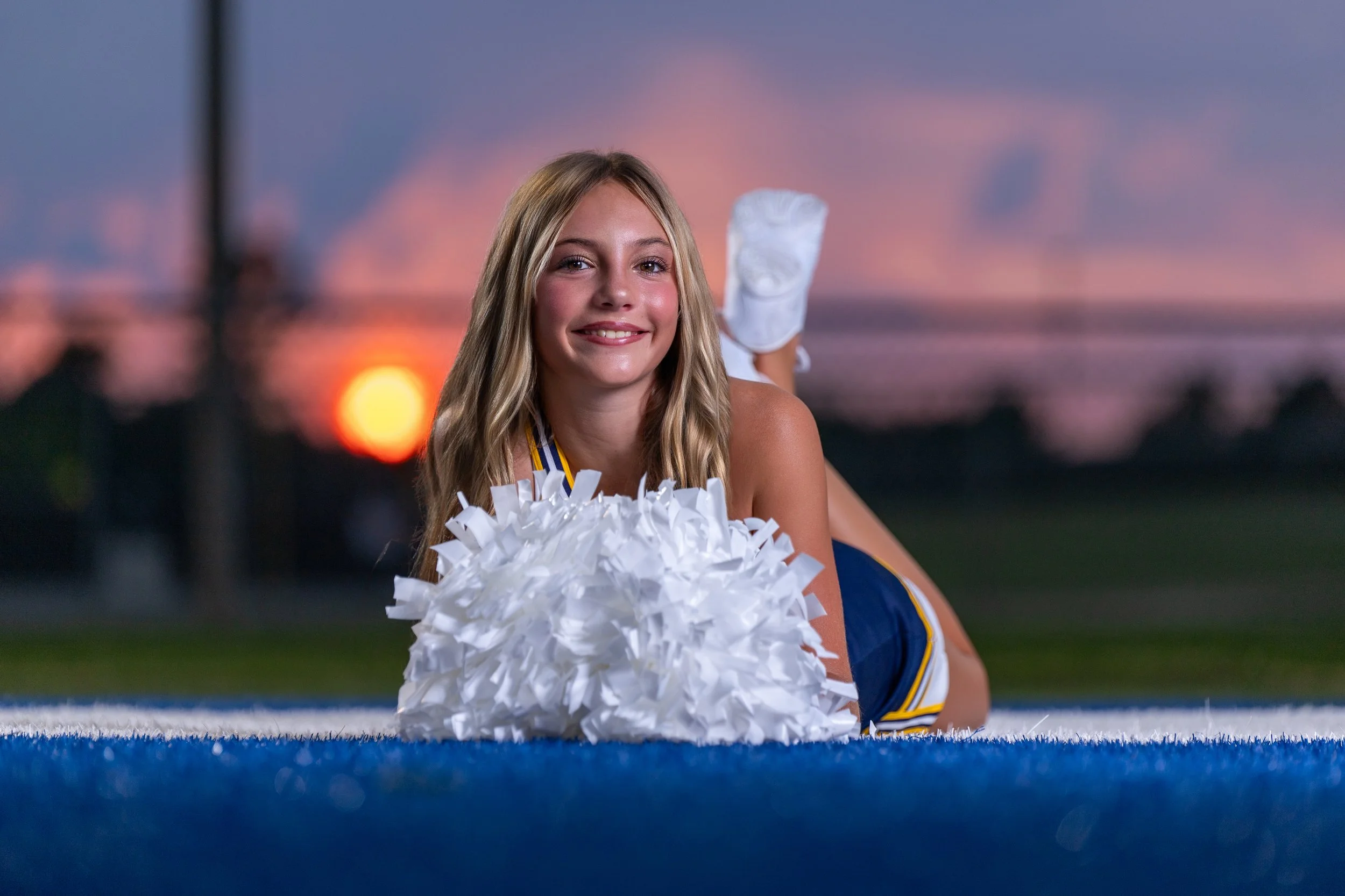 A cheerleader resting on a rail with white pom-poms, orange and pink sunset sky behind her.