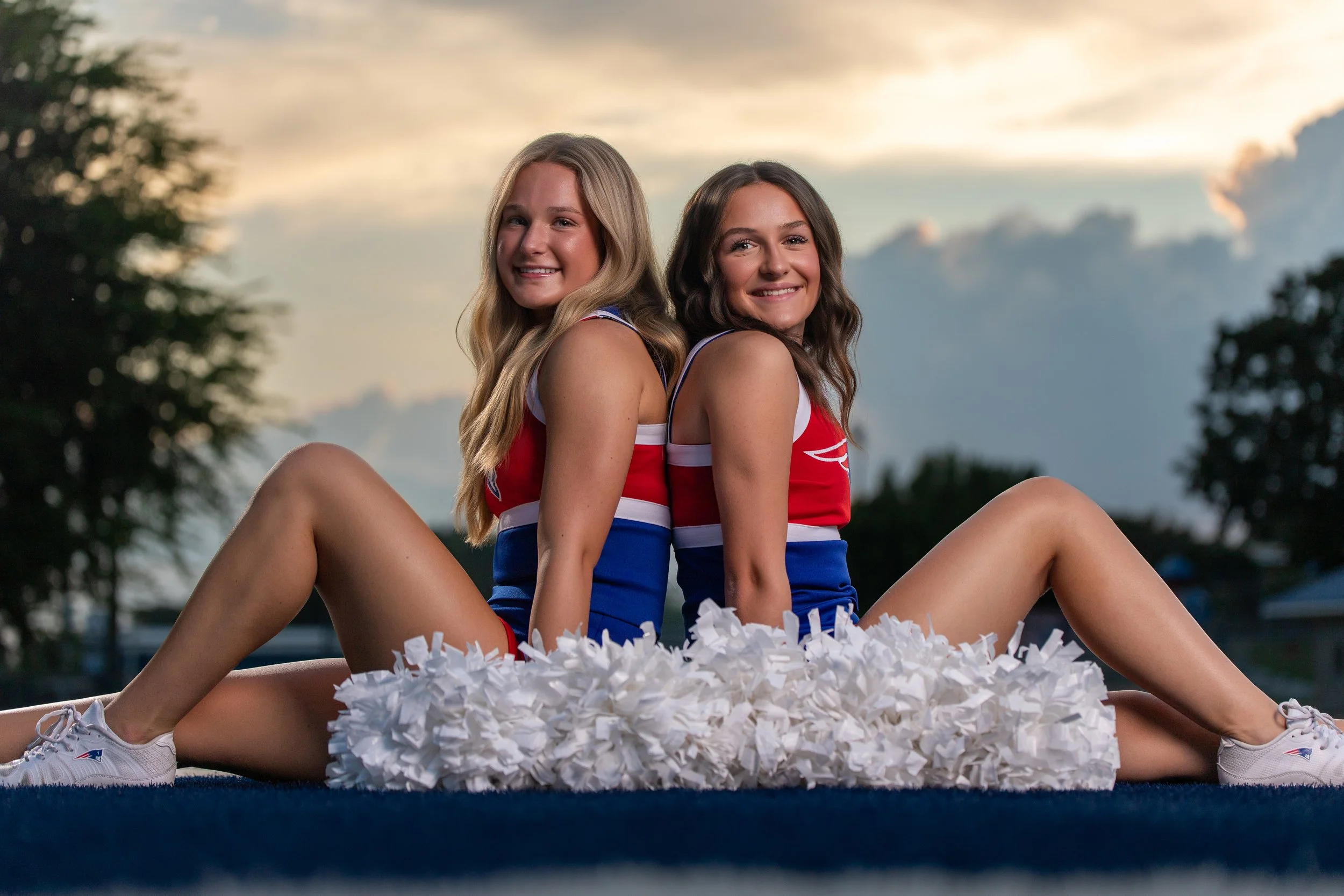 Two cheerleaders seated back to back with white pom-poms, sunset clouds behind them.