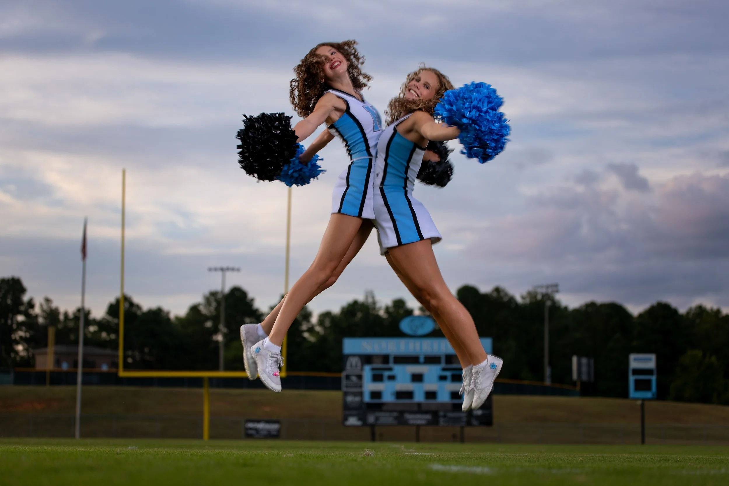Two cheerleaders jumping side by side with pom-poms raised, football field and scoreboard behind them at dusk.