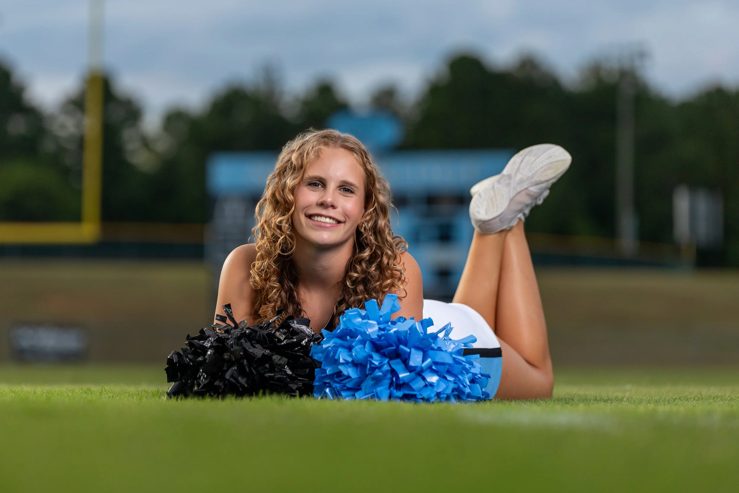 A cheerleader lying on the grass propped on her elbows with blue and black pom-poms.