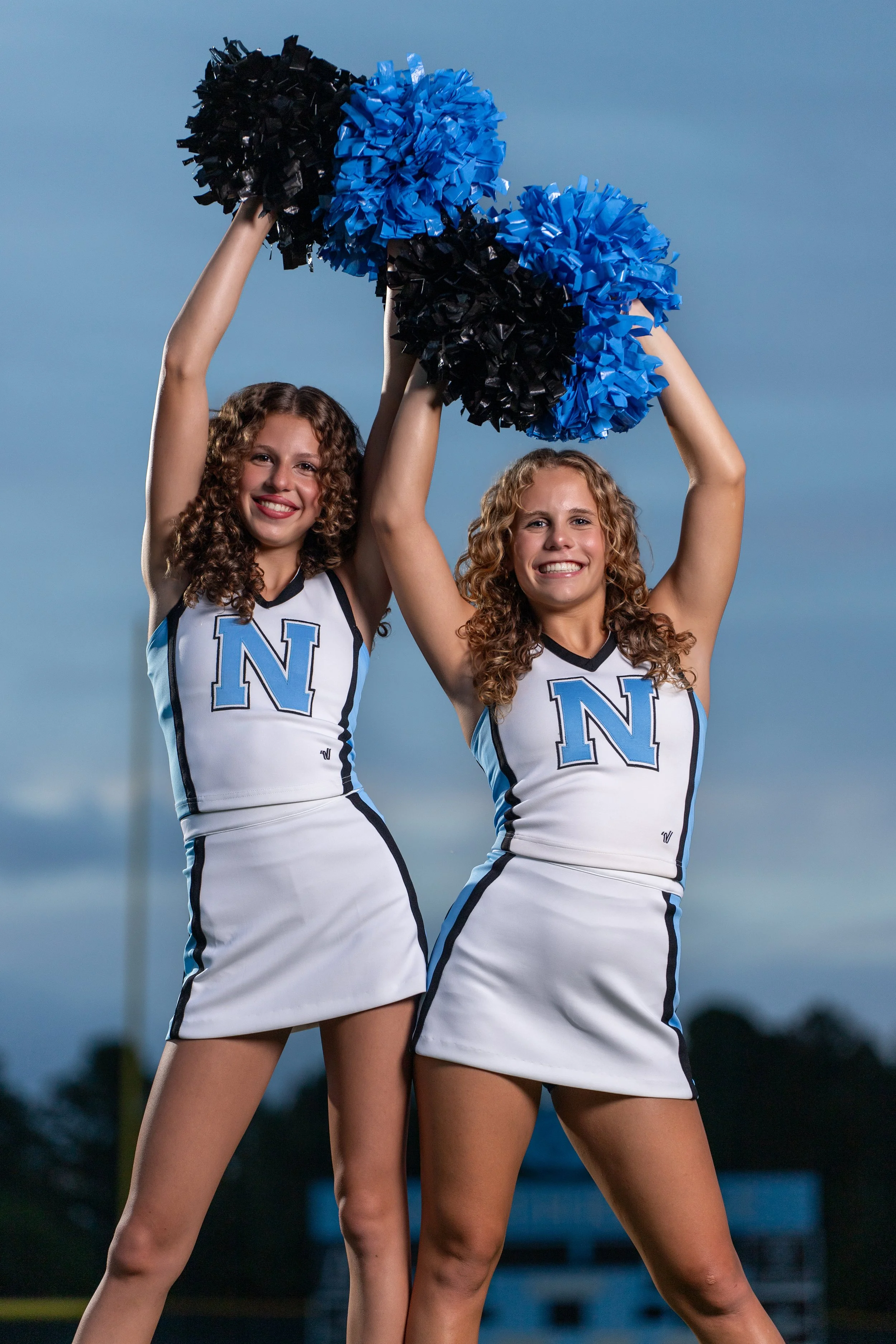 Two Northridge cheerleaders standing side by side with blue and black pom-poms raised overhead at dusk.