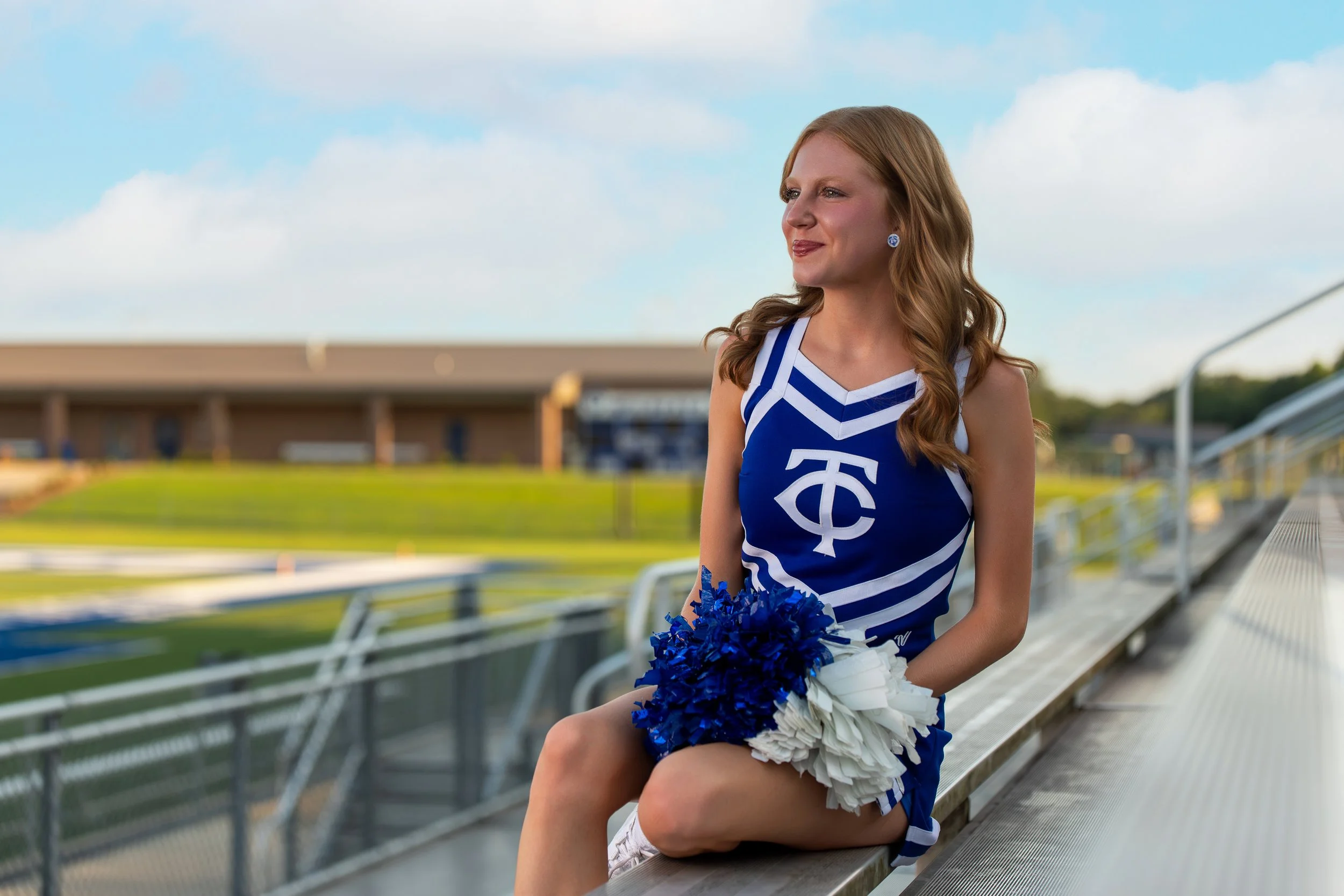 A cheerleader in a Tuscaloosa County uniform seated on stadium bleachers with pom-poms.